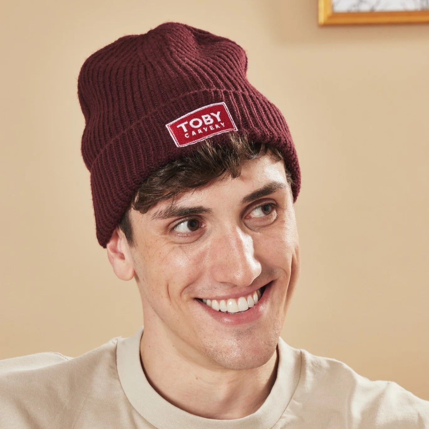 Person wearing a burgundy beanie with a Toby Carvery logo against a beige background