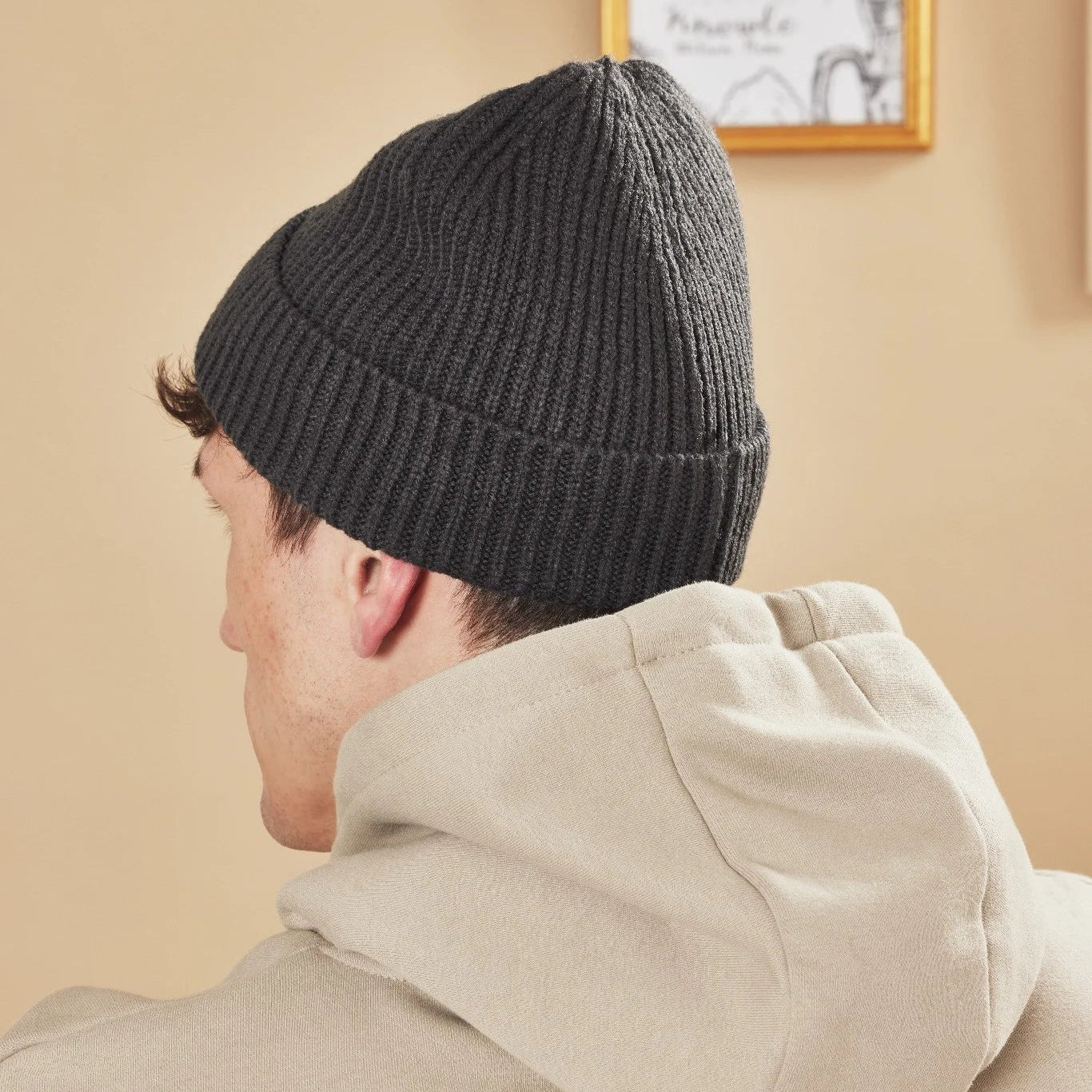 Person wearing a graphite knit beanie and beige jacket against a beige wall.
