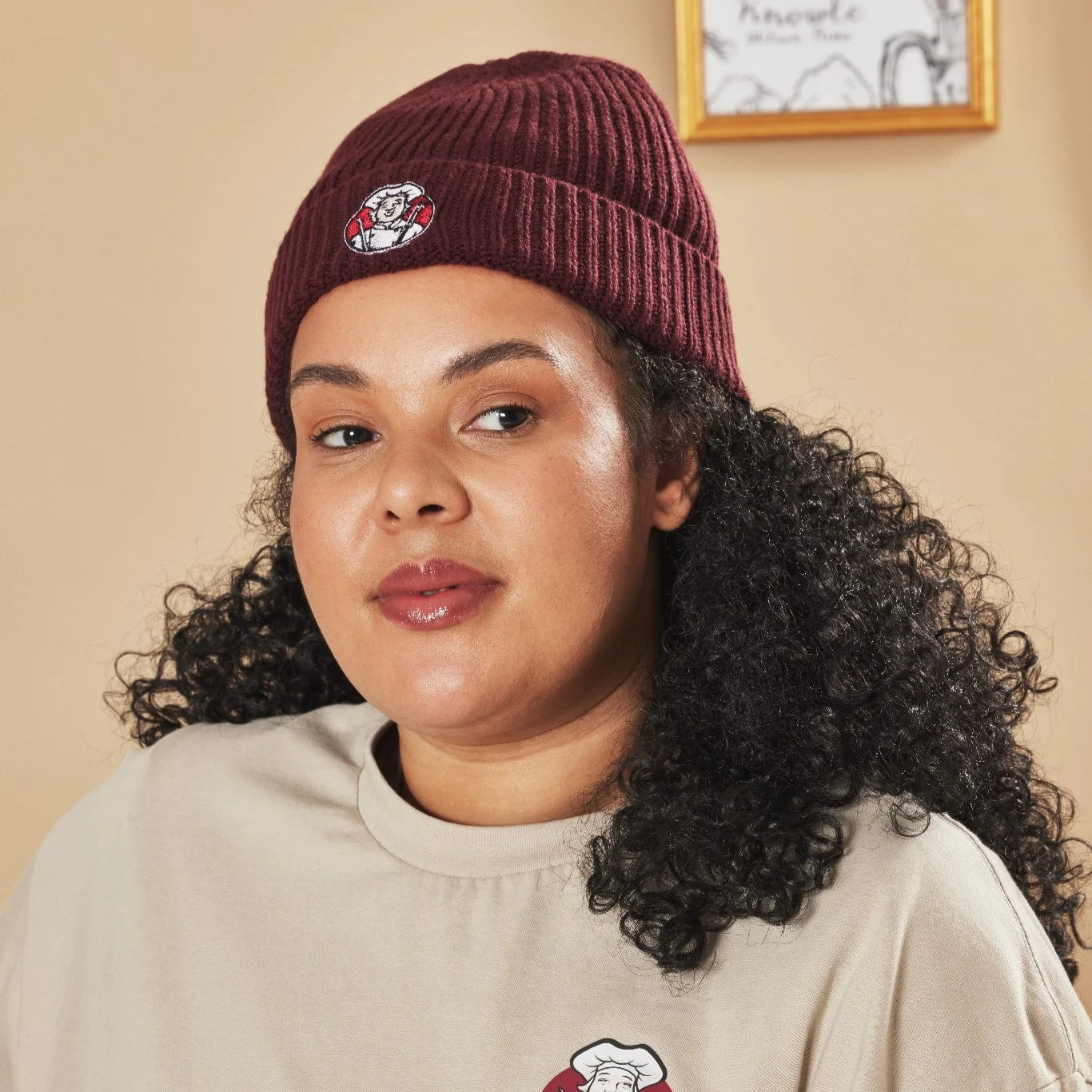 Woman wearing a Burgundy beanie and beige sweatshirt with Toby Carvery logos, standing against a beige wall.