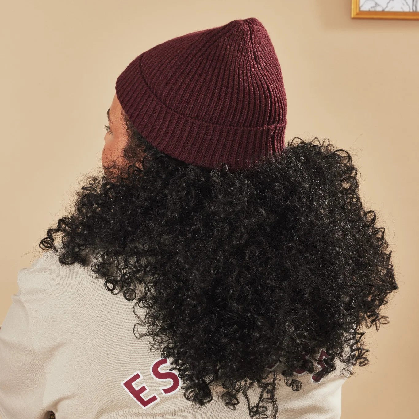 Person wearing a burgundy beanie with curly hair, standing against a beige wall.