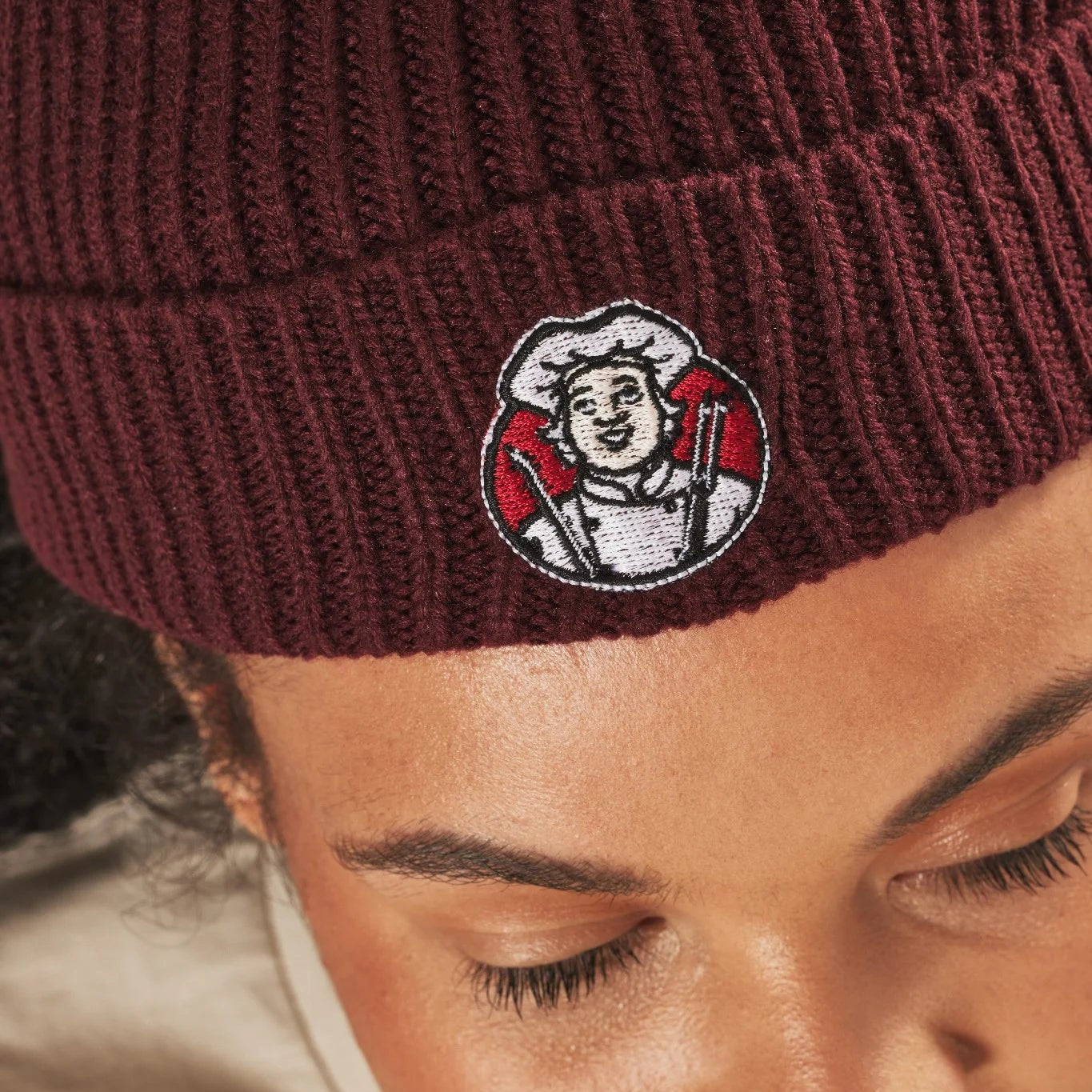 Close-up of a burgundy knit beanie with an embroidered Toby Carvery logo on a person's head.