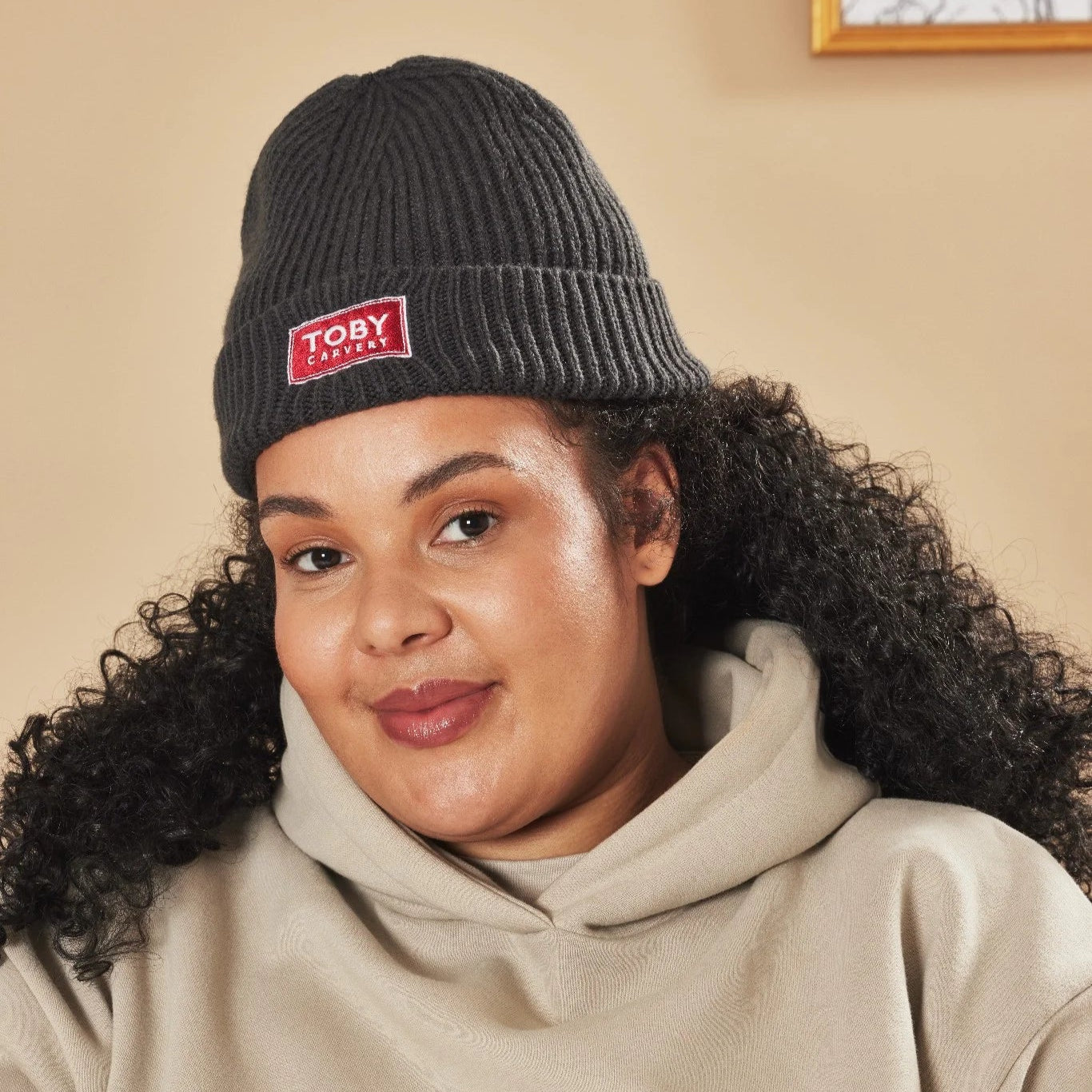 Person wearing a graphite beanie with a Toby Carvery logo and beige hoodie indoors.