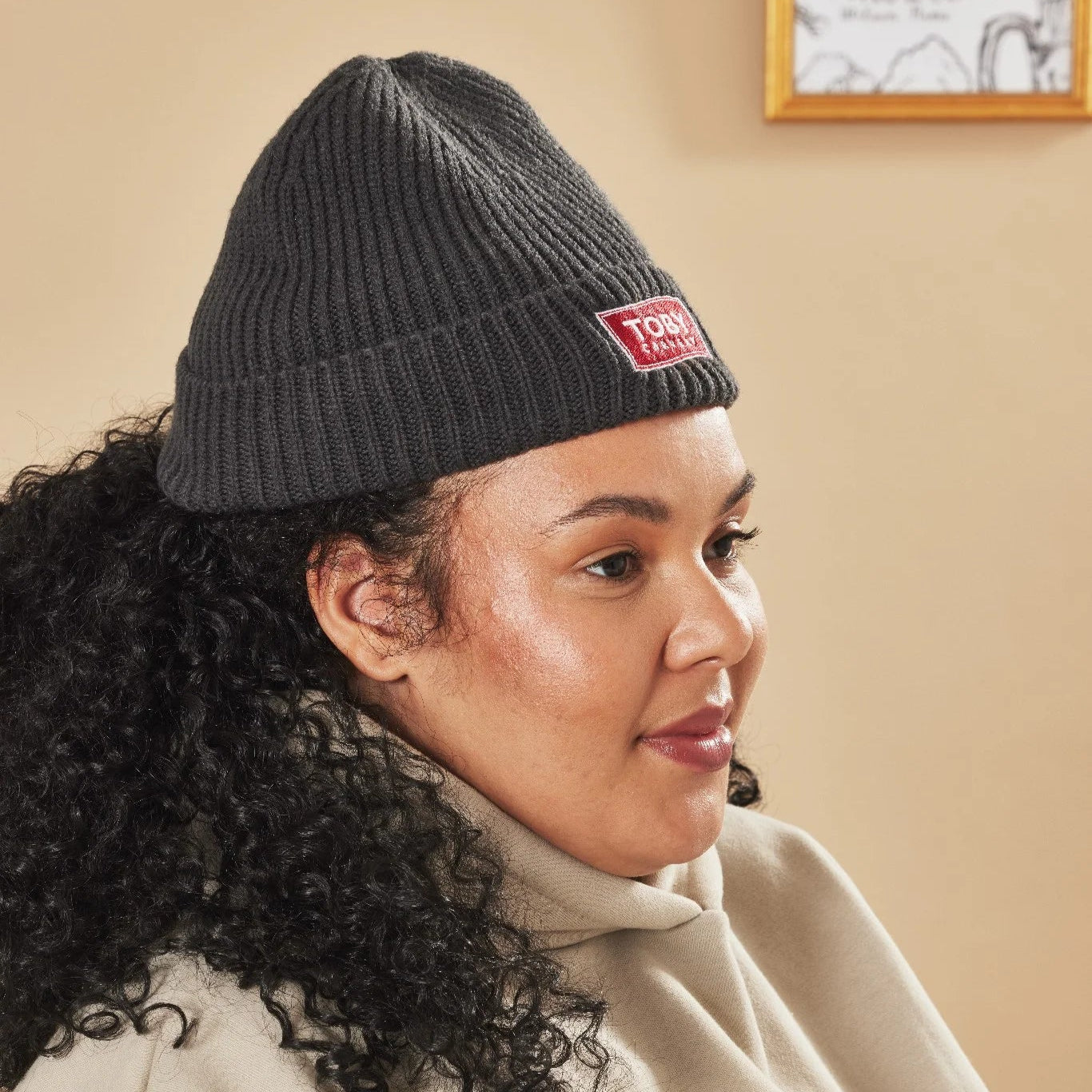 Woman wearing a graphite knit beanie with a Toby Carvery logo, sitting indoors.
