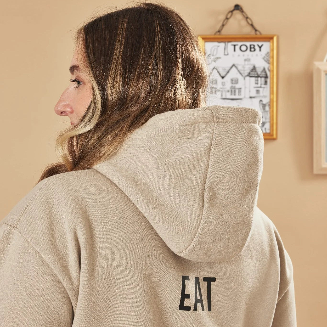 Person wearing a natural stone hoodie with 'EAT' text, standing in a room with a framed picture on the wall.