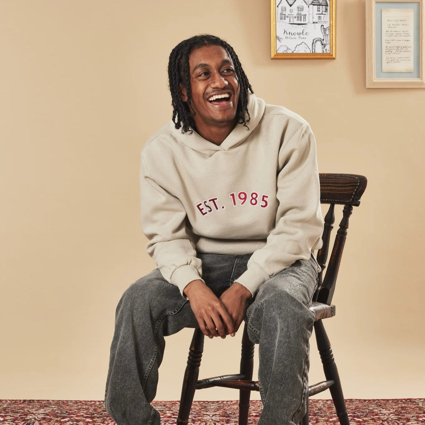 Person wearing a natural stone hoodie with 'EST. 1985' text, sitting on a wooden chair.