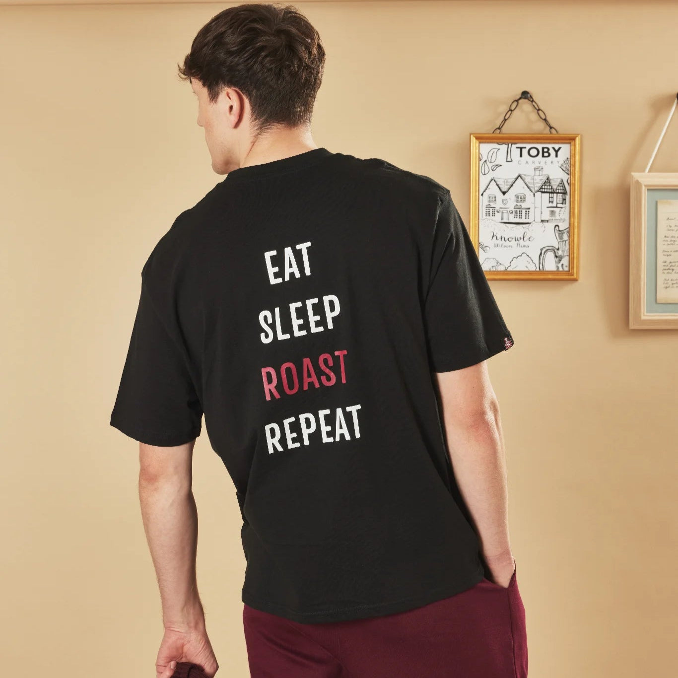 Person wearing a black t-shirt with 'EAT SLEEP ROAST REPEAT' text on the back, standing against a beige wall.