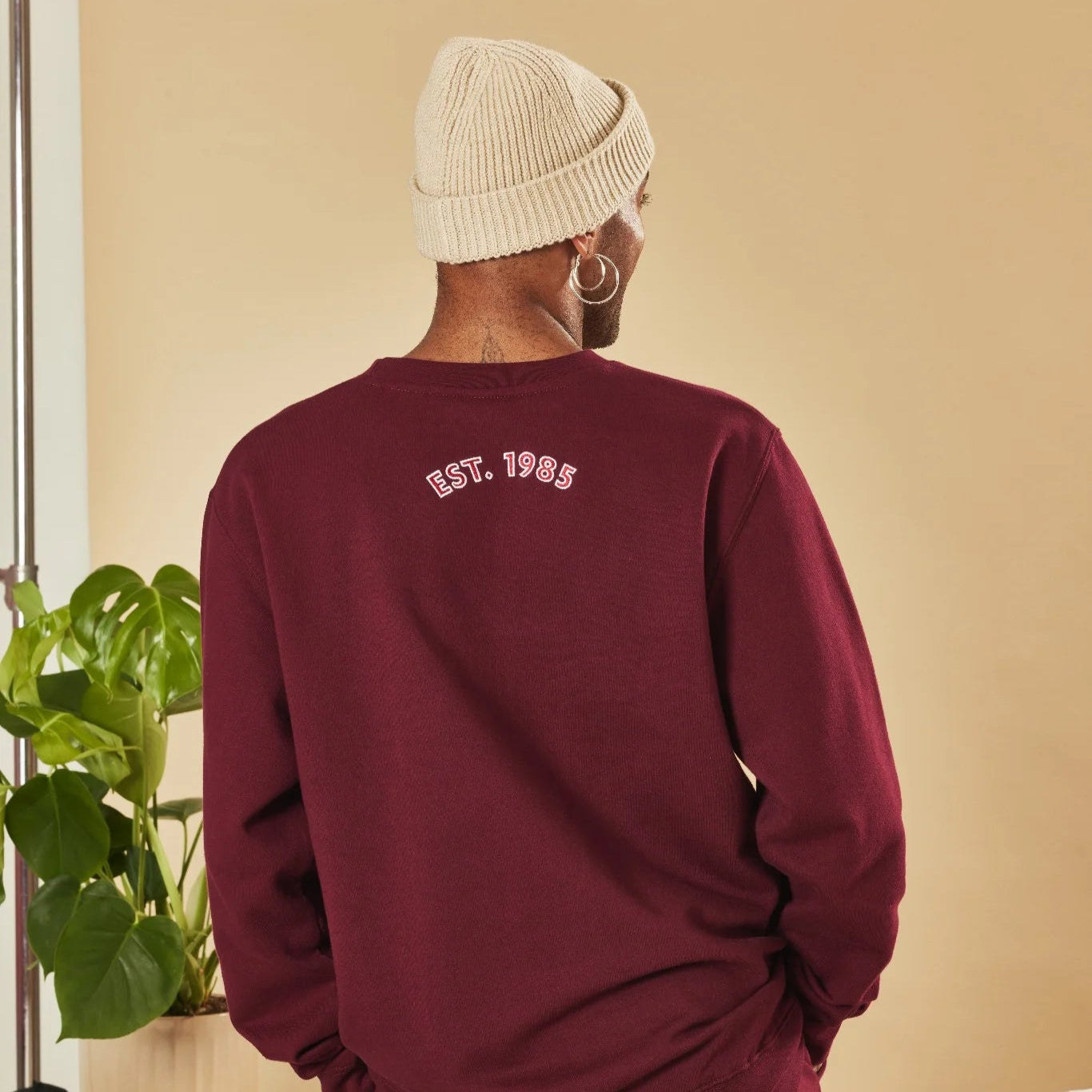 Person wearing a burgundy sweatshirt with 'EST. 1985' text, standing indoors with a plant in the corner.