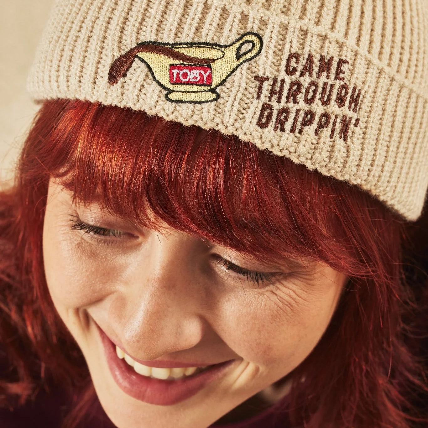 Person wearing an oatmeal knit beanie with a logo and text, smiling.