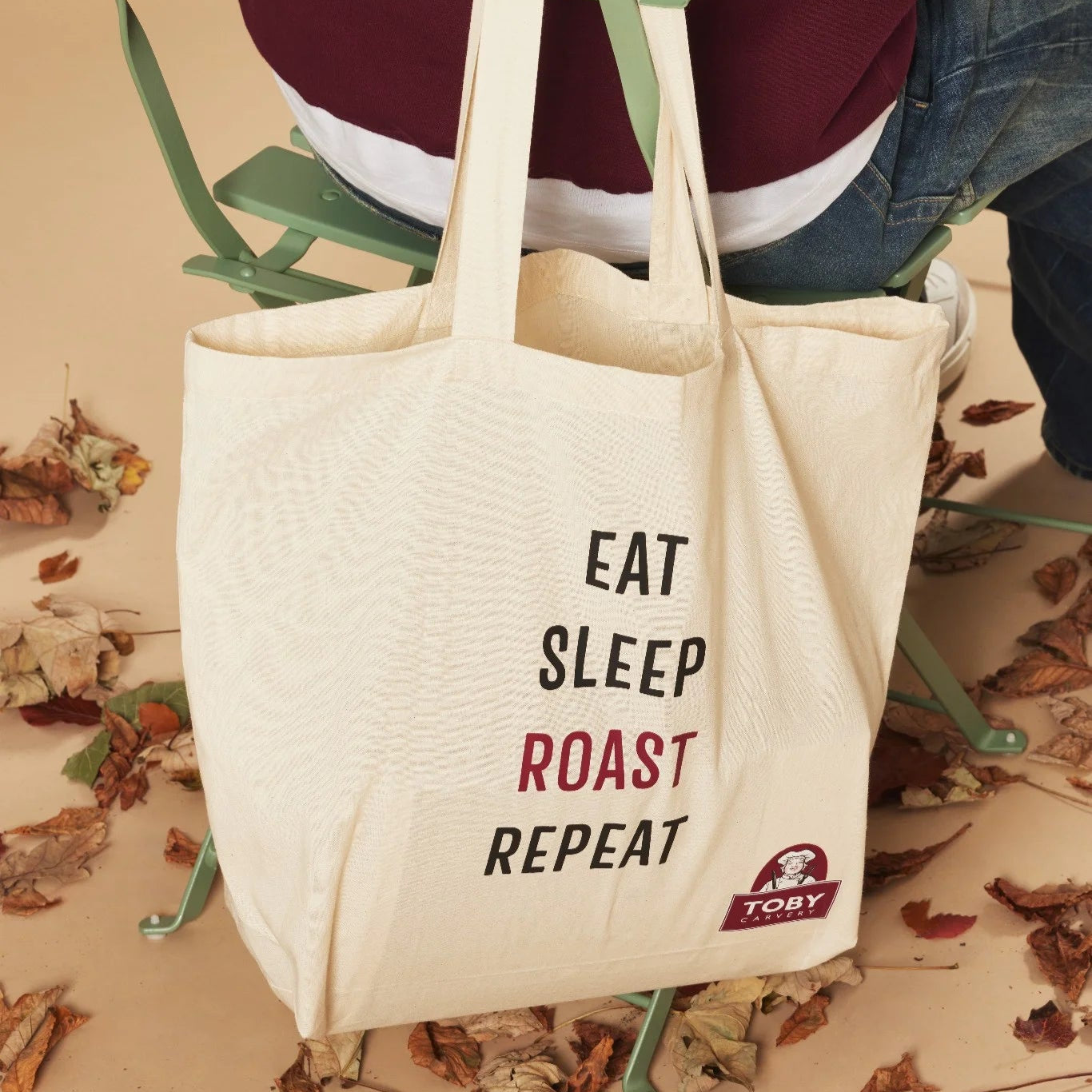 Tote bag with 'Eat Sleep Roast Repeat' text on a person sitting on a chair with leaves on the ground.