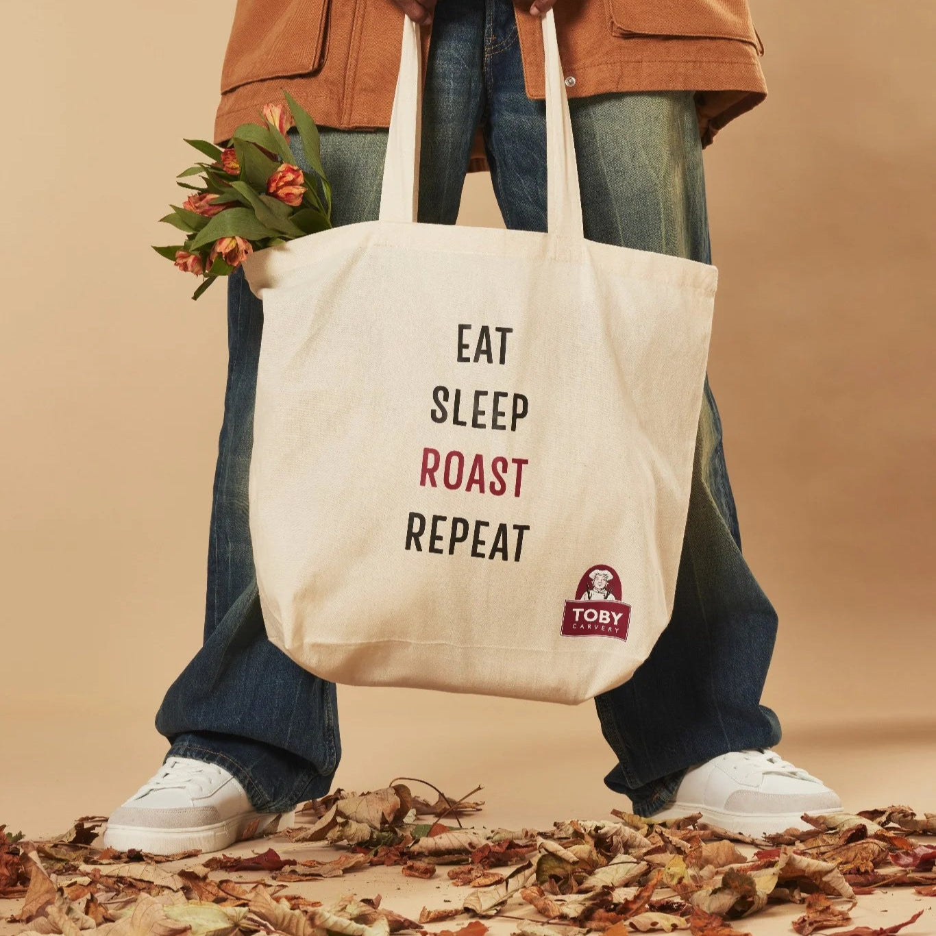 Person holding a tote bag with 'Eat Sleep Roast Repeat' text and a Toby Carvery logo, standing on leaves against a beige background.