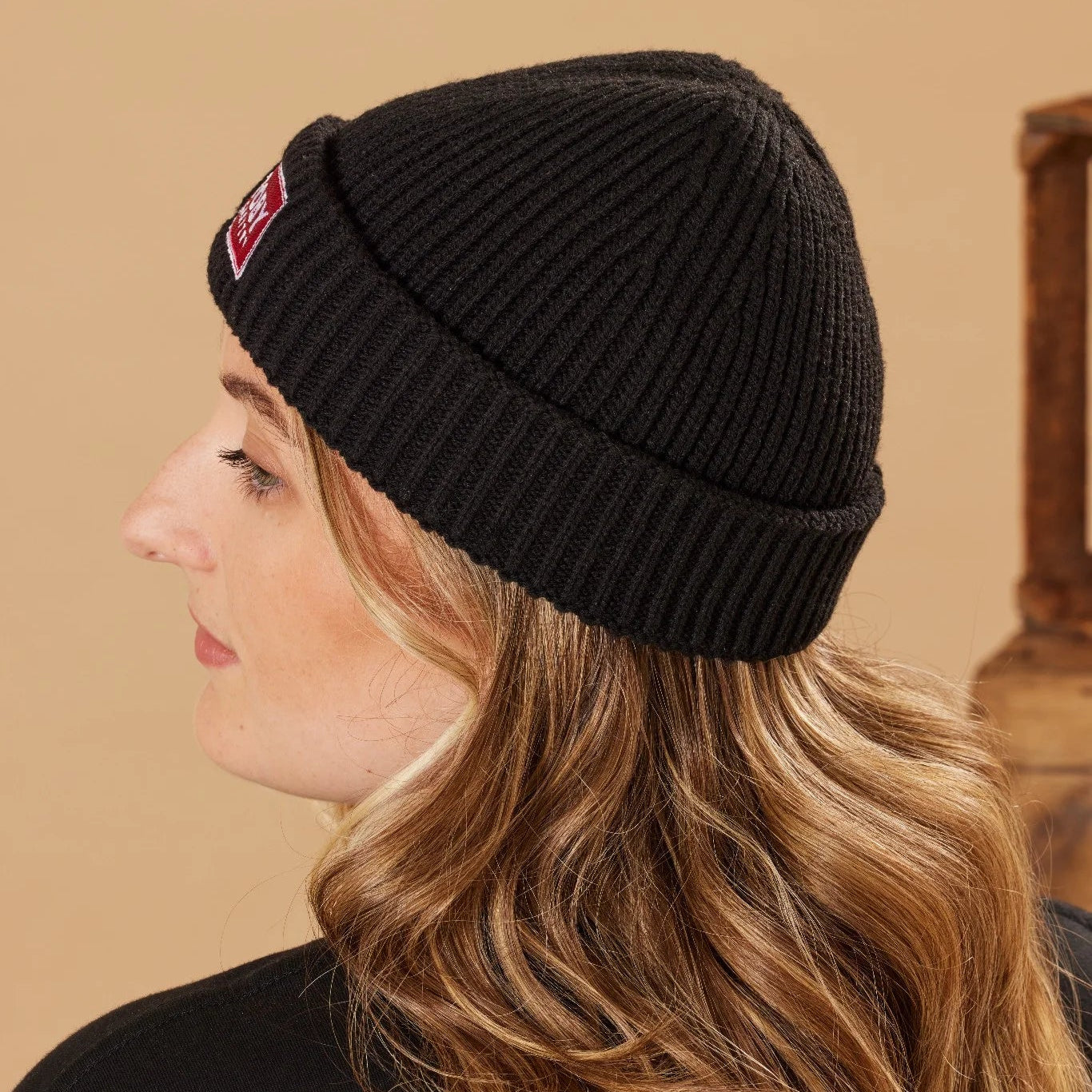 Person wearing a black beanie with a red patch on a beige background