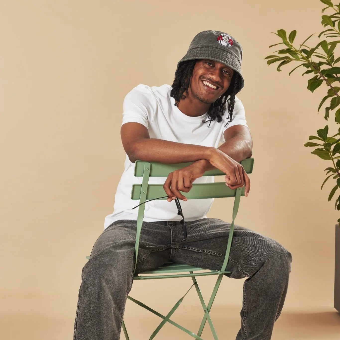 Man sitting on a green chair wearing a white t-shirt, dark jeans, and a graphite bucket hat with a plant in the background.