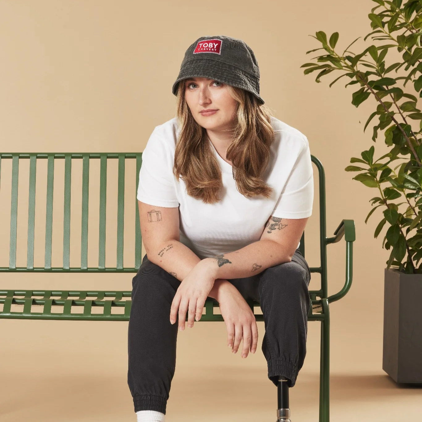 Person sitting on a green bench wearing a white t-shirt and black cap with a logo, against a beige background.