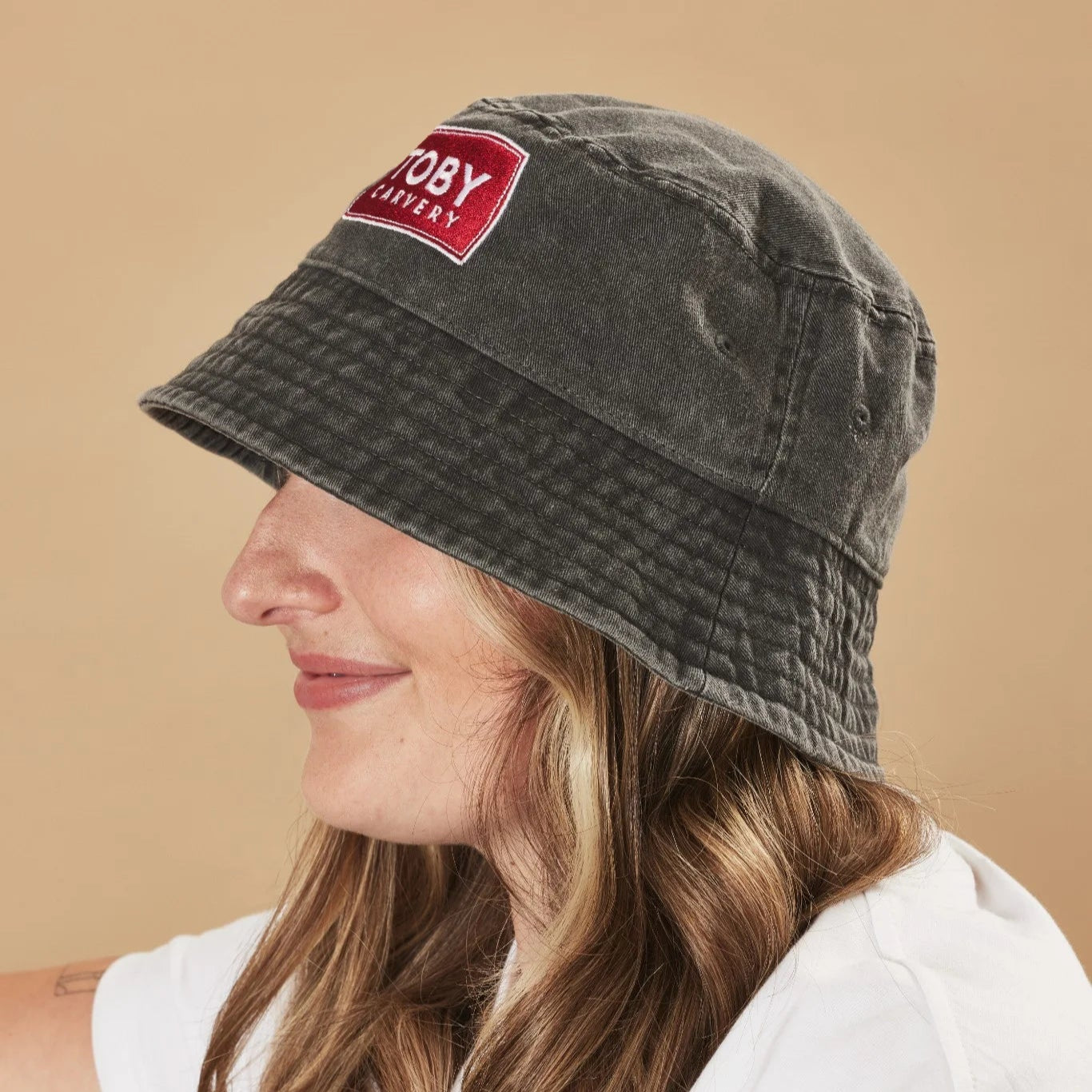 Person wearing a graphite bucket hat with a Toby Carvery logo on a beige background