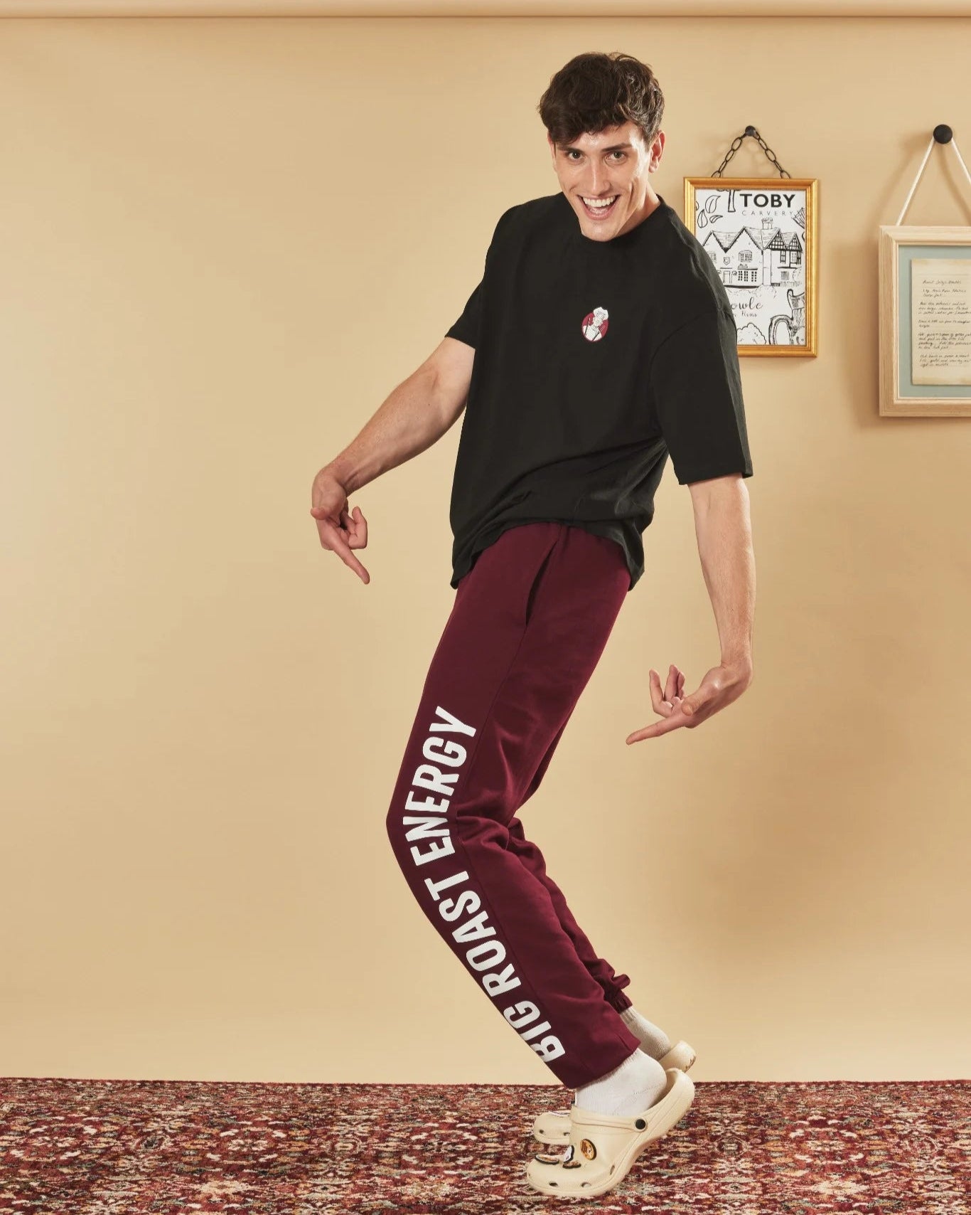 Person wearing a black t-shirt and burgundy joggers with text, standing against a beige wall.