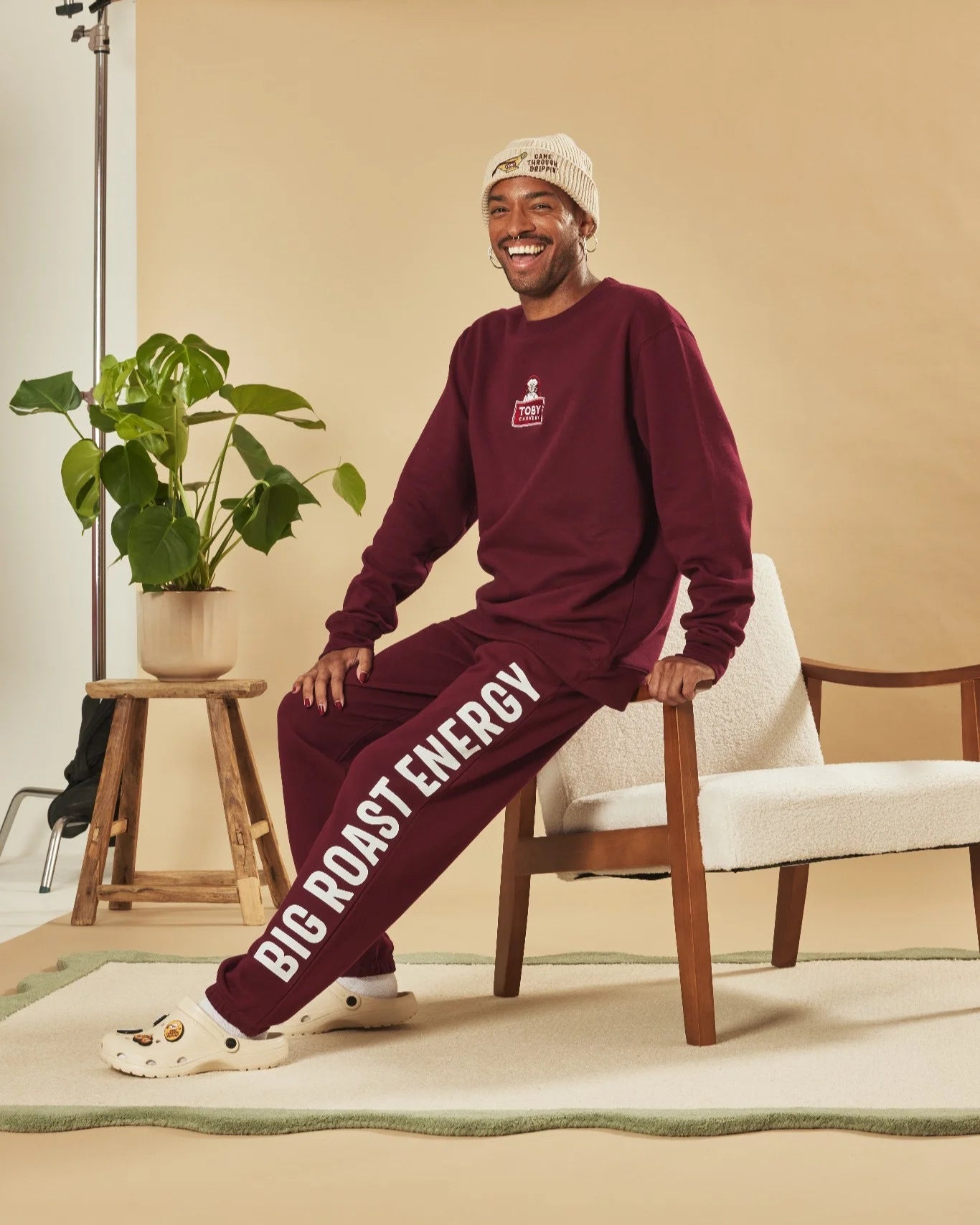 Man wearing a burgundy tracksuit with 'Big Roast Energy' text, sitting on a chair in a room with a plant and chair.