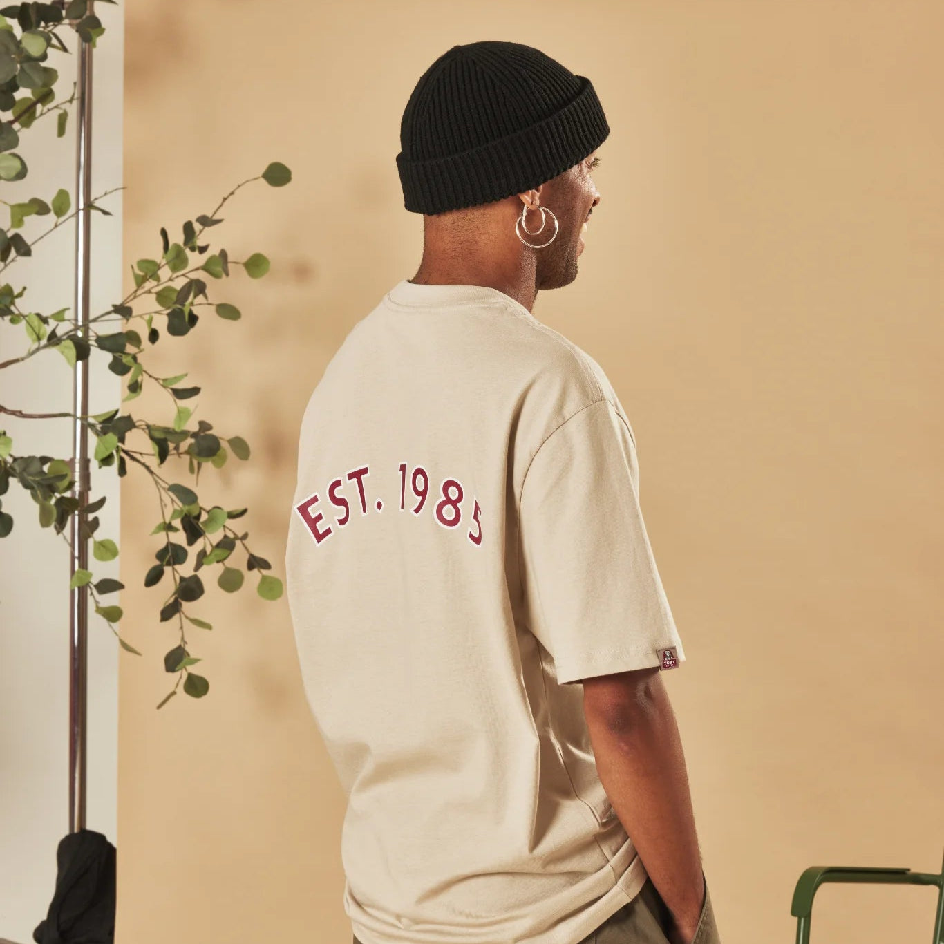 Person wearing a natural stone t-shirt with 'EST. 1985' text, standing against a beige wall with a plant on the left.