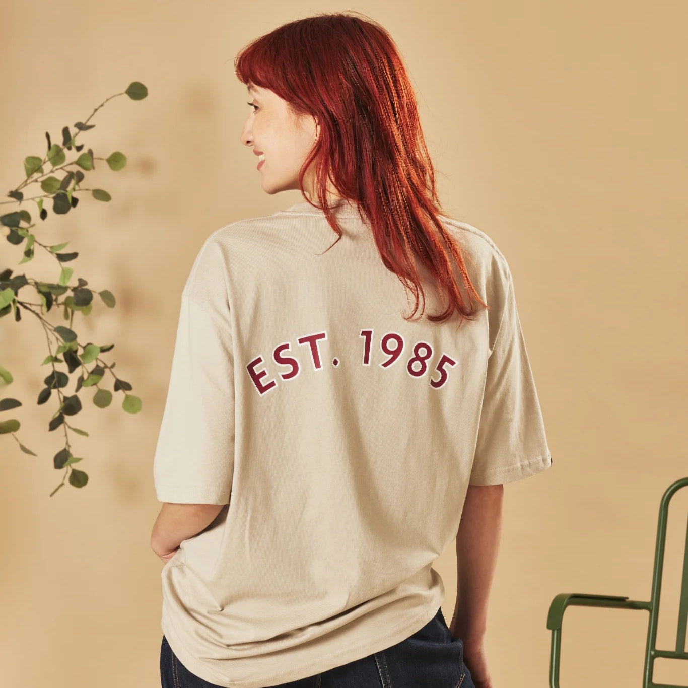 Person wearing a natural stone t-shirt with 'EST. 1985' text on the back, standing against a beige wall with a plant.