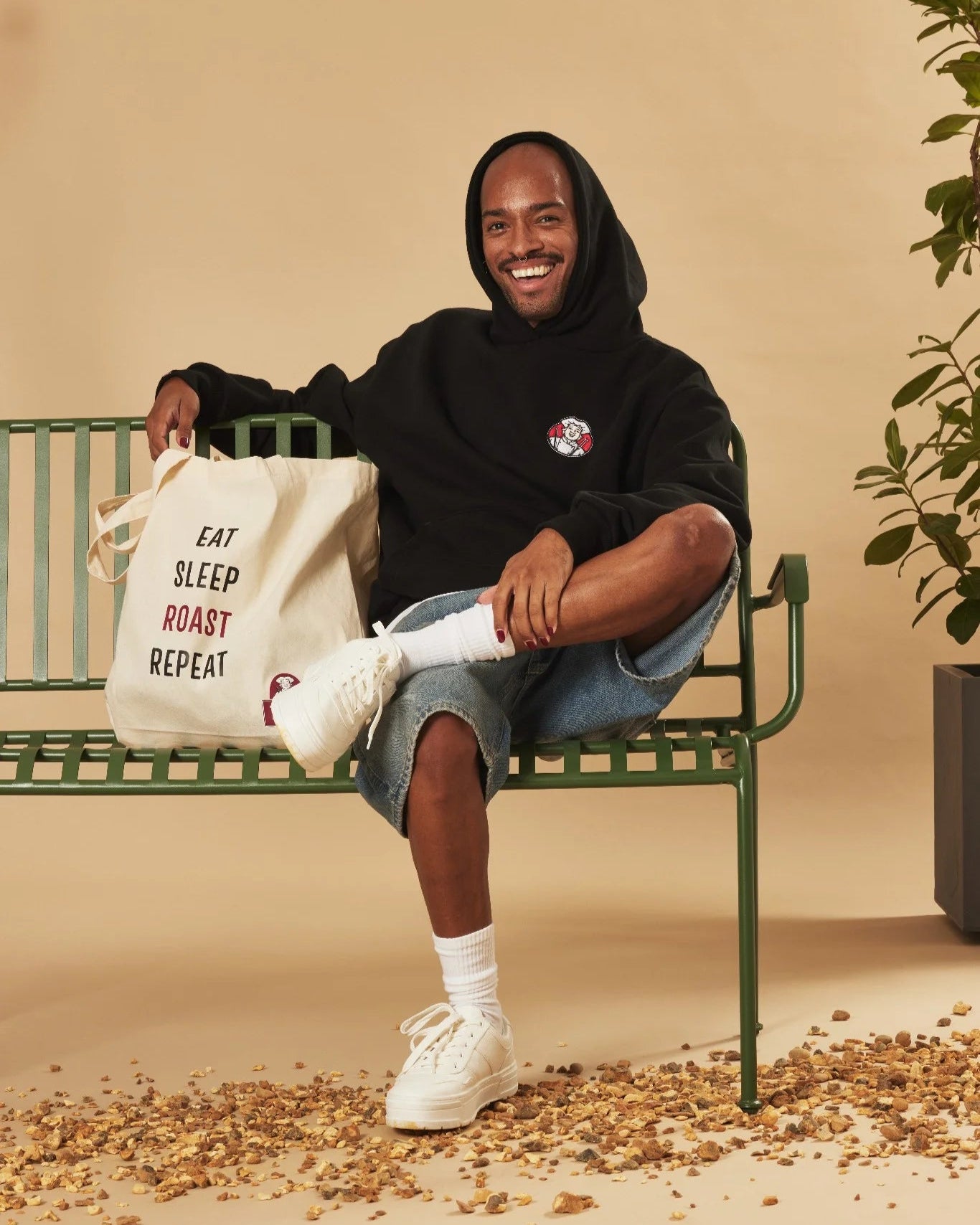 Person sitting on a bench holding a tote bag with text, wearing a black hoodie and white trainers.