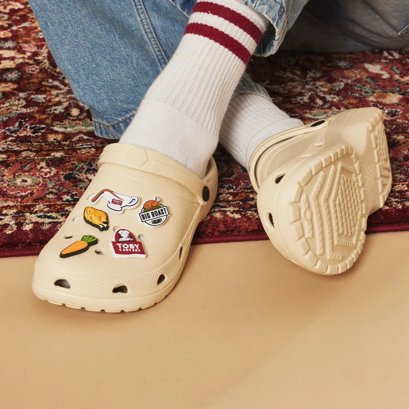 Beige clogs with Toby Carvery Charms worn by a person on a patterned rug.