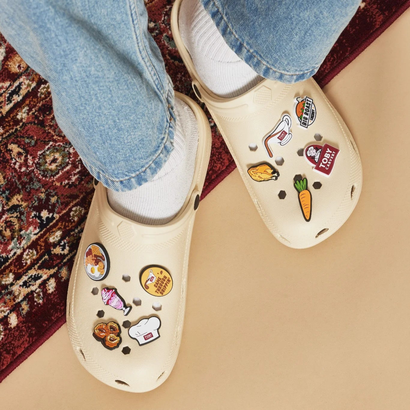 Beige clogs with Toby Carvery shoe charms worn with blue jeans and white socks on a patterned rug.