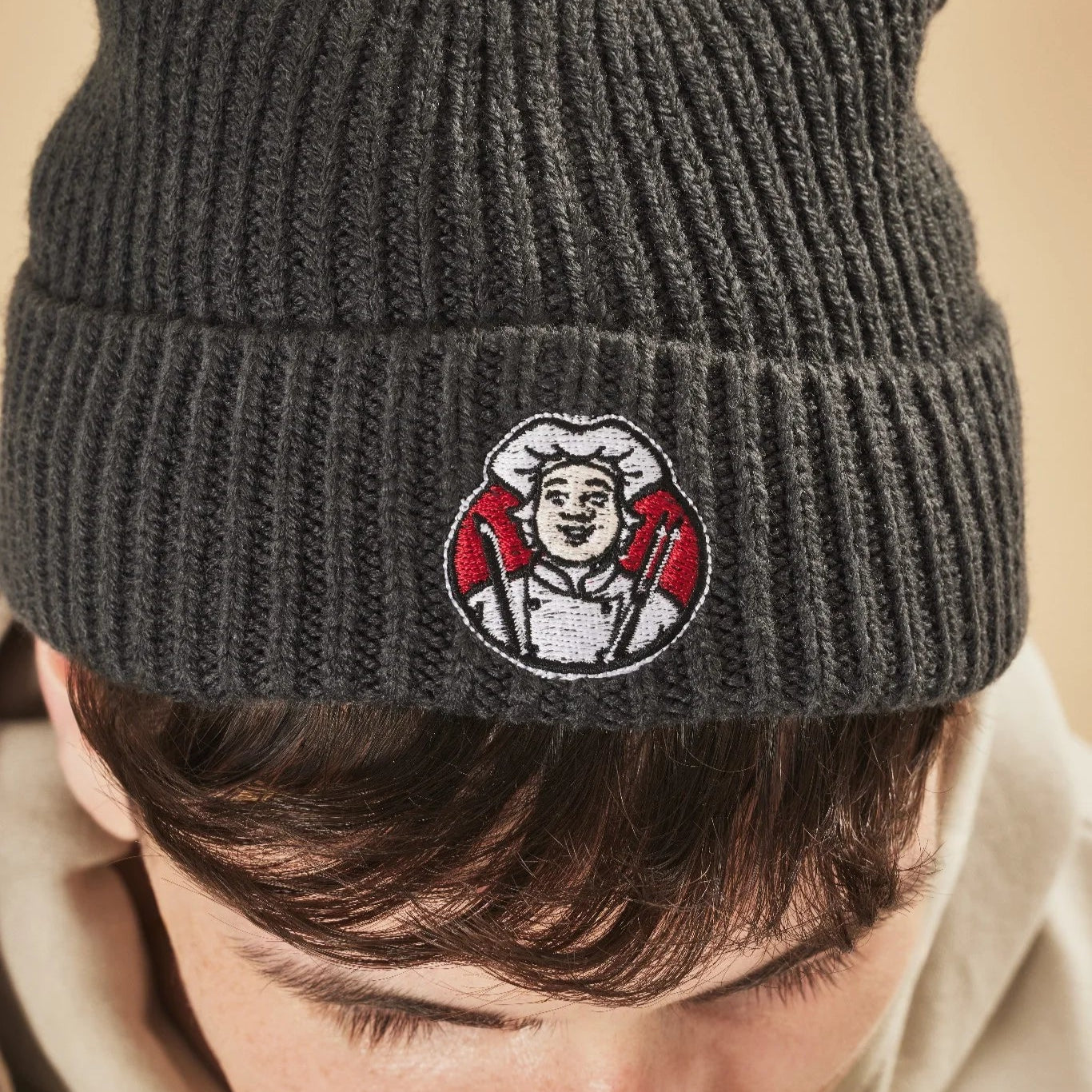 Graphite knit beanie with an embroidered Toby Carvery logo worn by a person, against a beige background