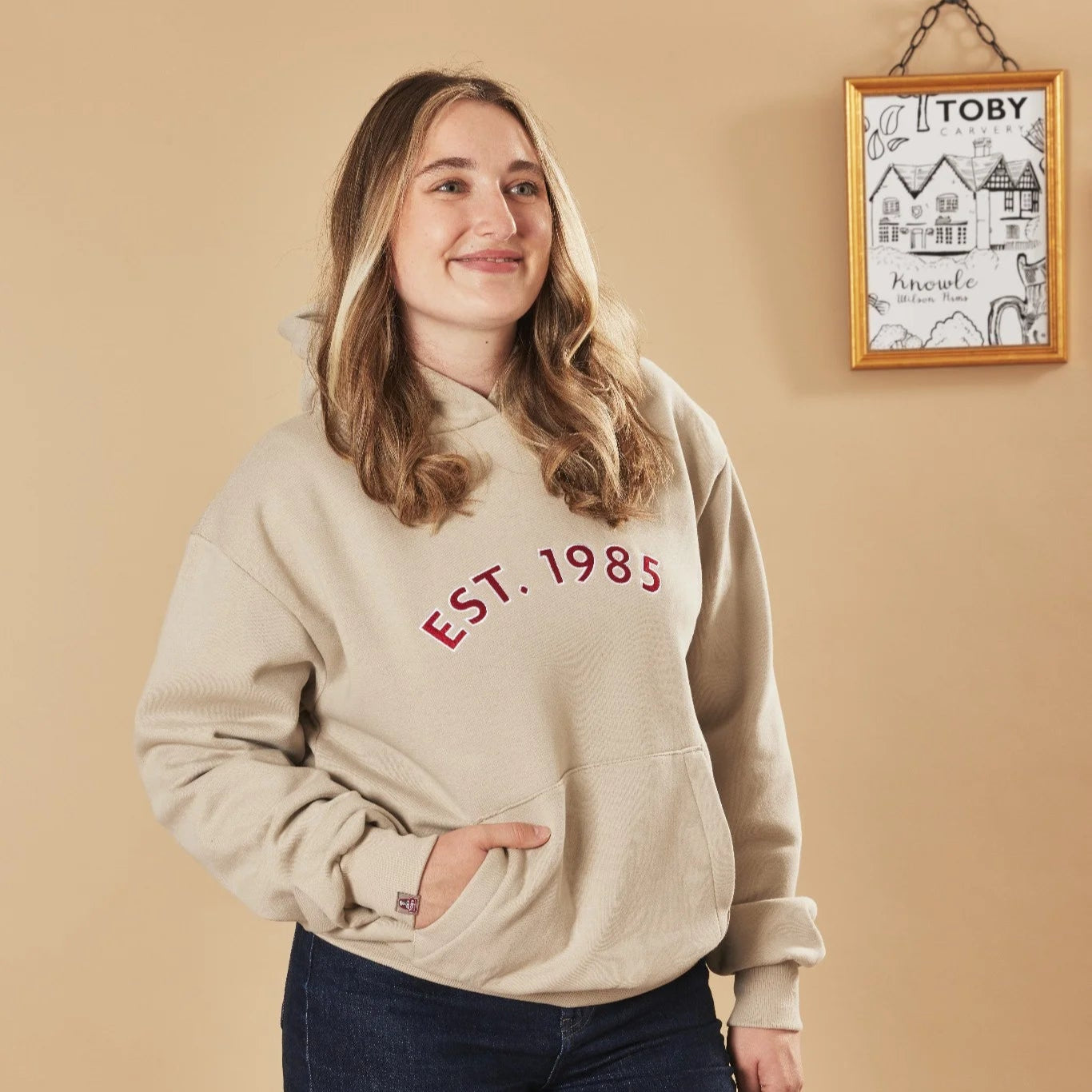 Woman wearing a natural stone hoodie with 'EST. 1985' text, standing against a beige wall with a framed picture.