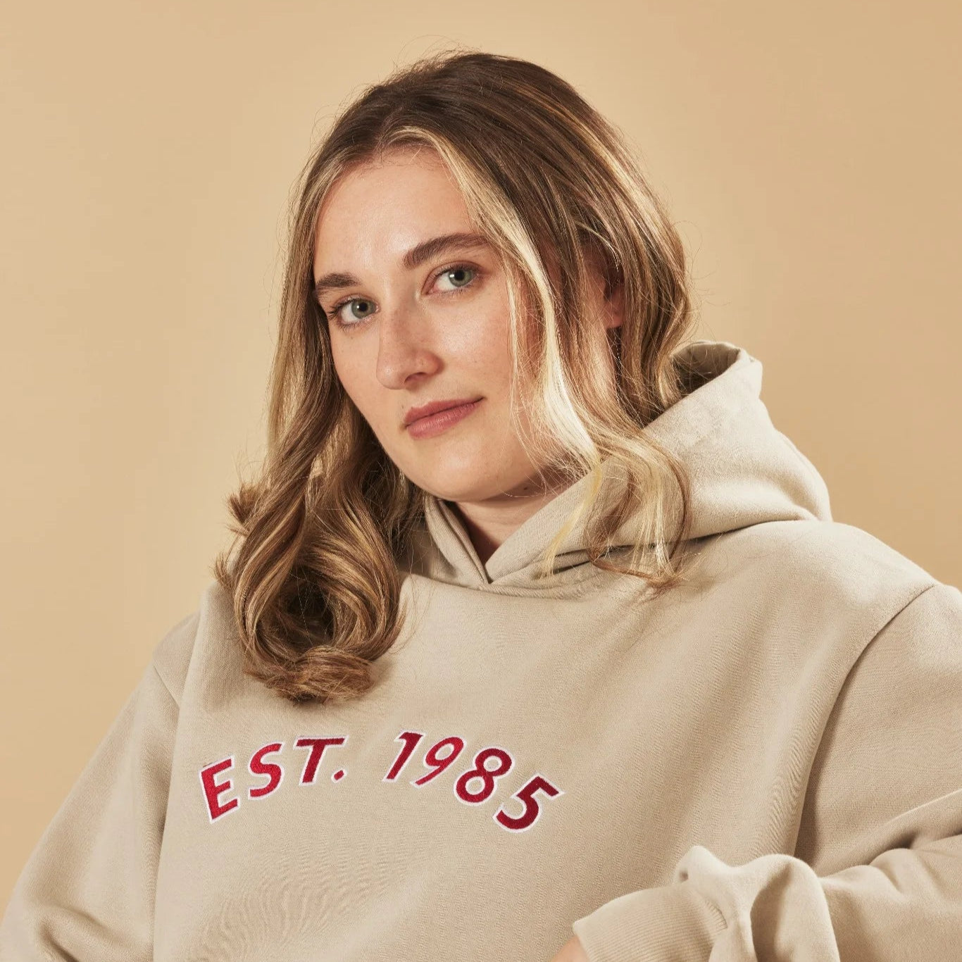 Woman wearing a natural stone hoodie with 'EST. 1985' text on a beige background