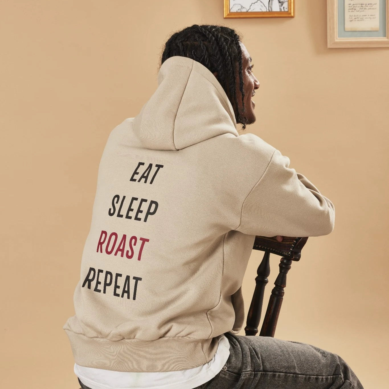 Person wearing a natural stone hoodie with 'Eat Sleep Roast Repeat' text, sitting on a chair.