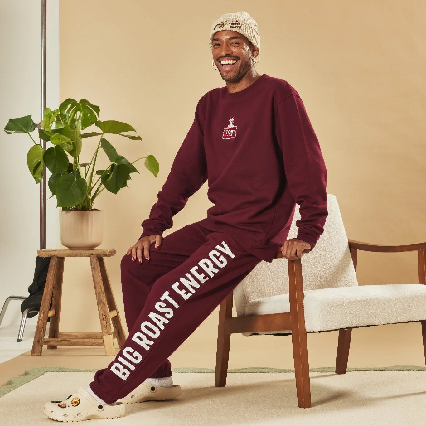 Man wearing a burgundy sweatshirt and joggers with text, sitting on a chair in a room with a plant and stool.