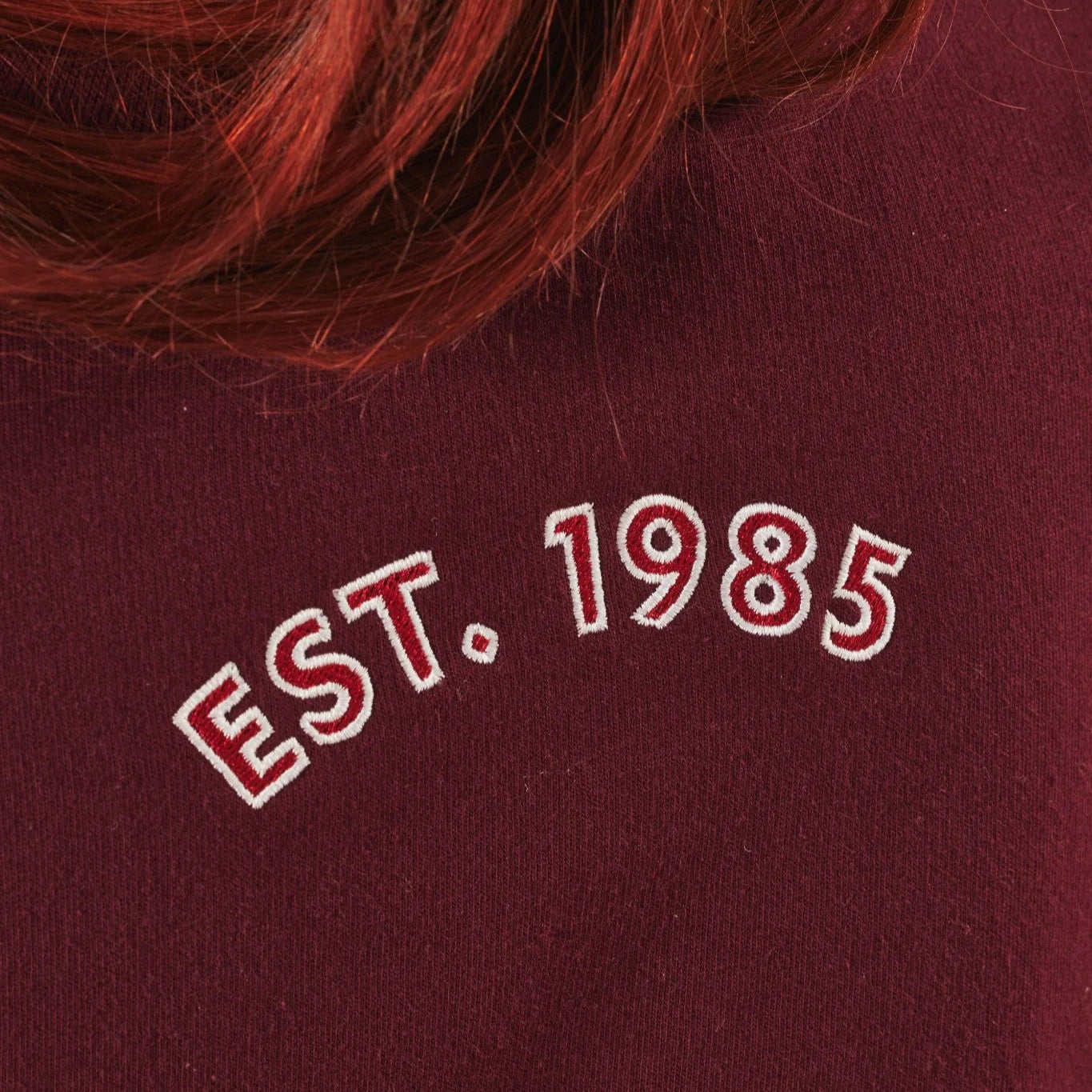 Burgundy sweatshirt with 'EST. 1985' text on a blurred background