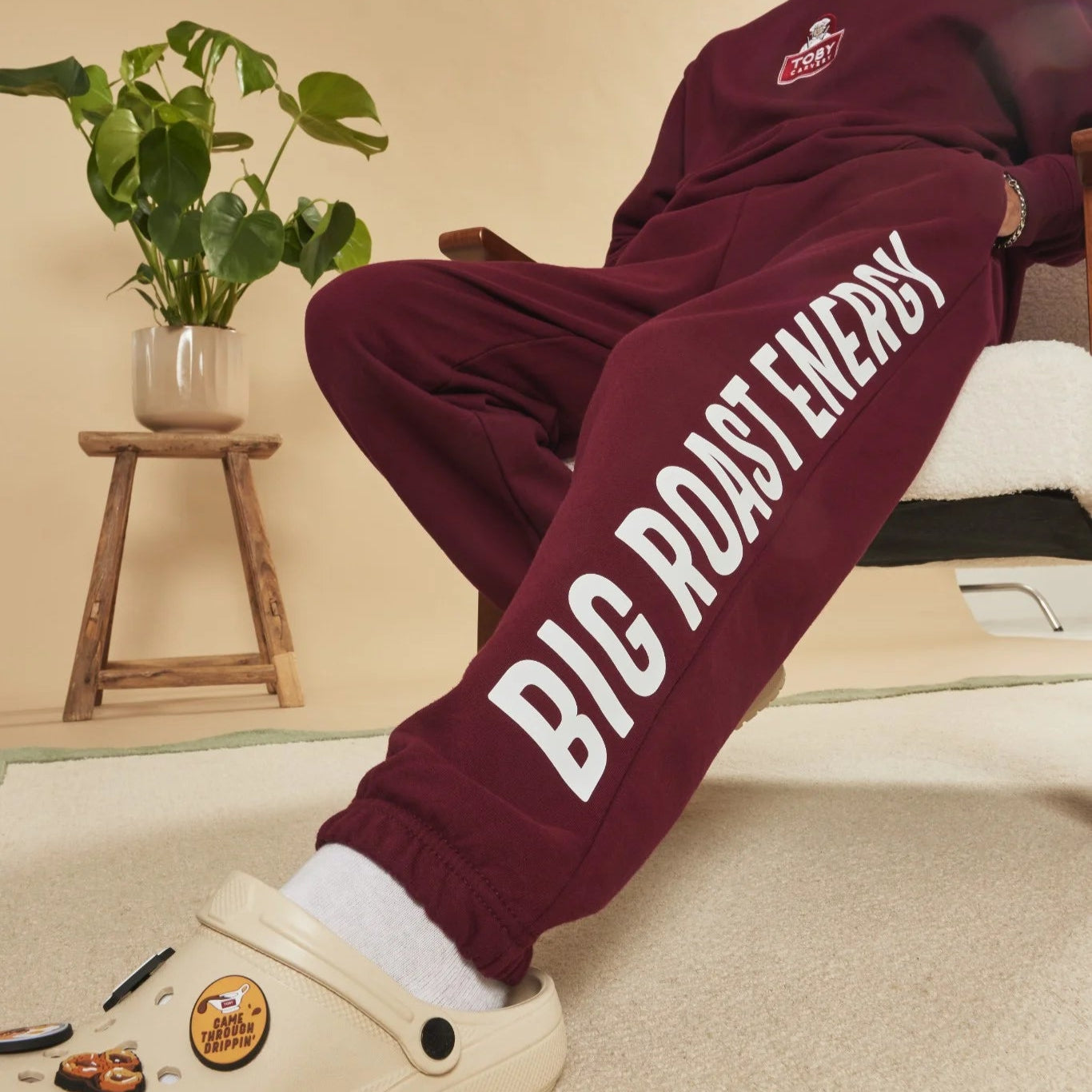 Burgundy joggers with 'BIG ROAST ENERGY' text worn by a person, sitting on a chair.