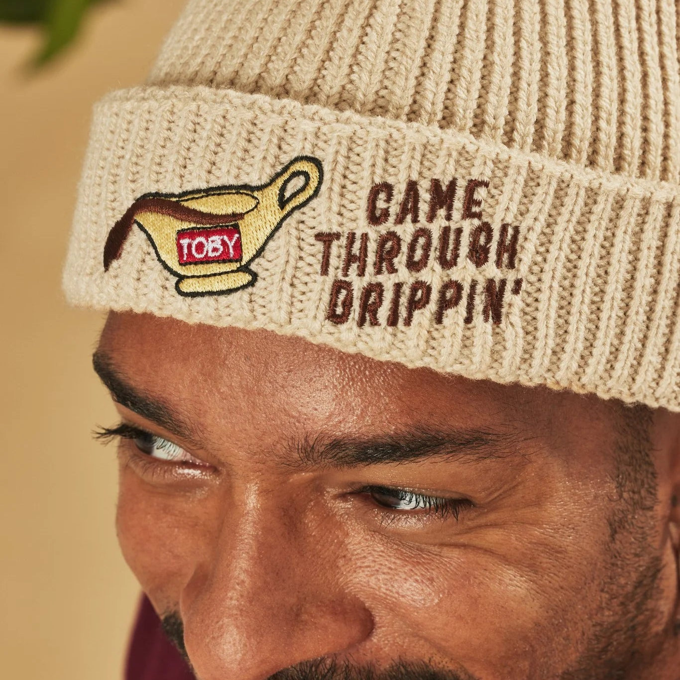 Man wearing an oatmeal beanie with text and logo on a beige background