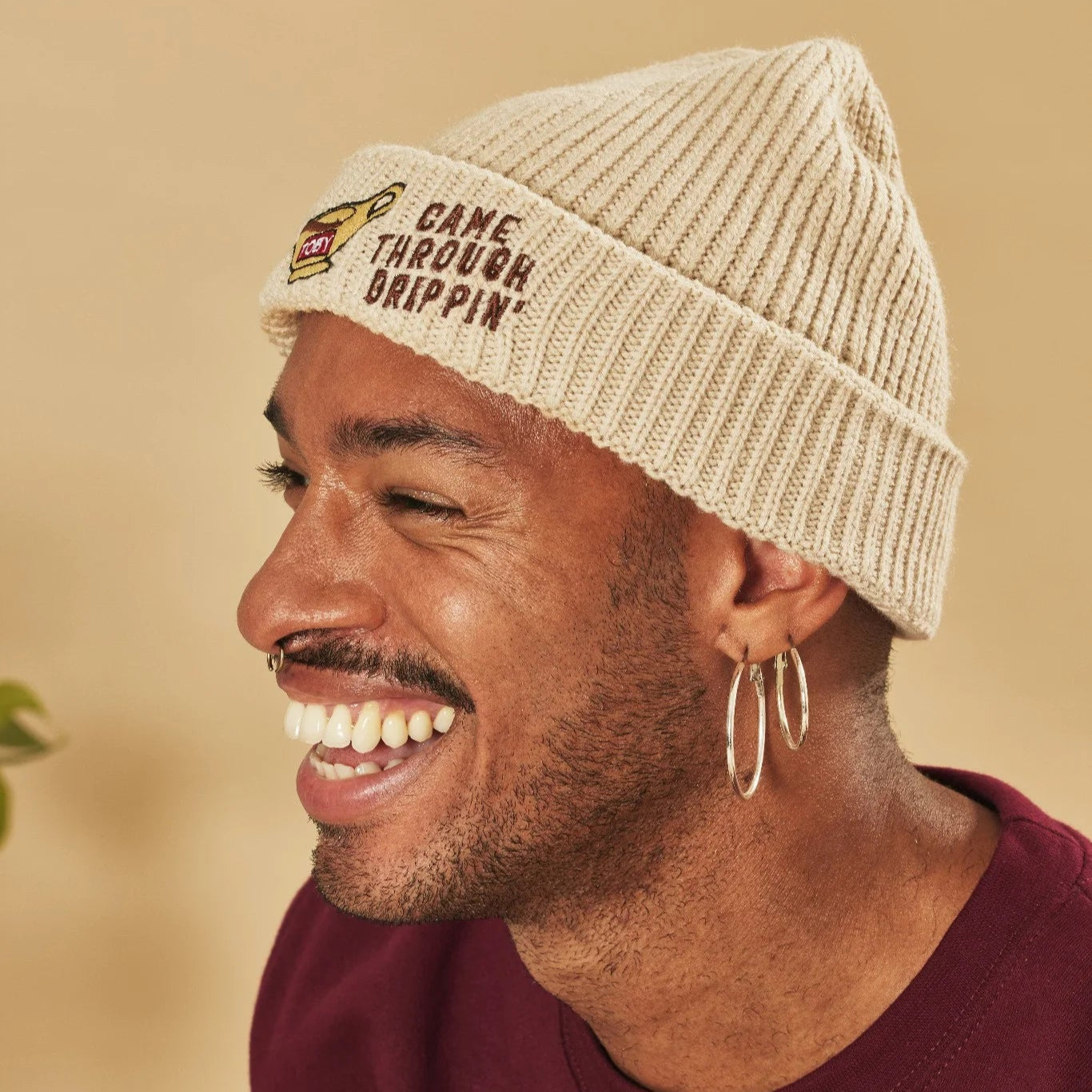 Man wearing an oatmeal beanie with text, smiling against a blurred background