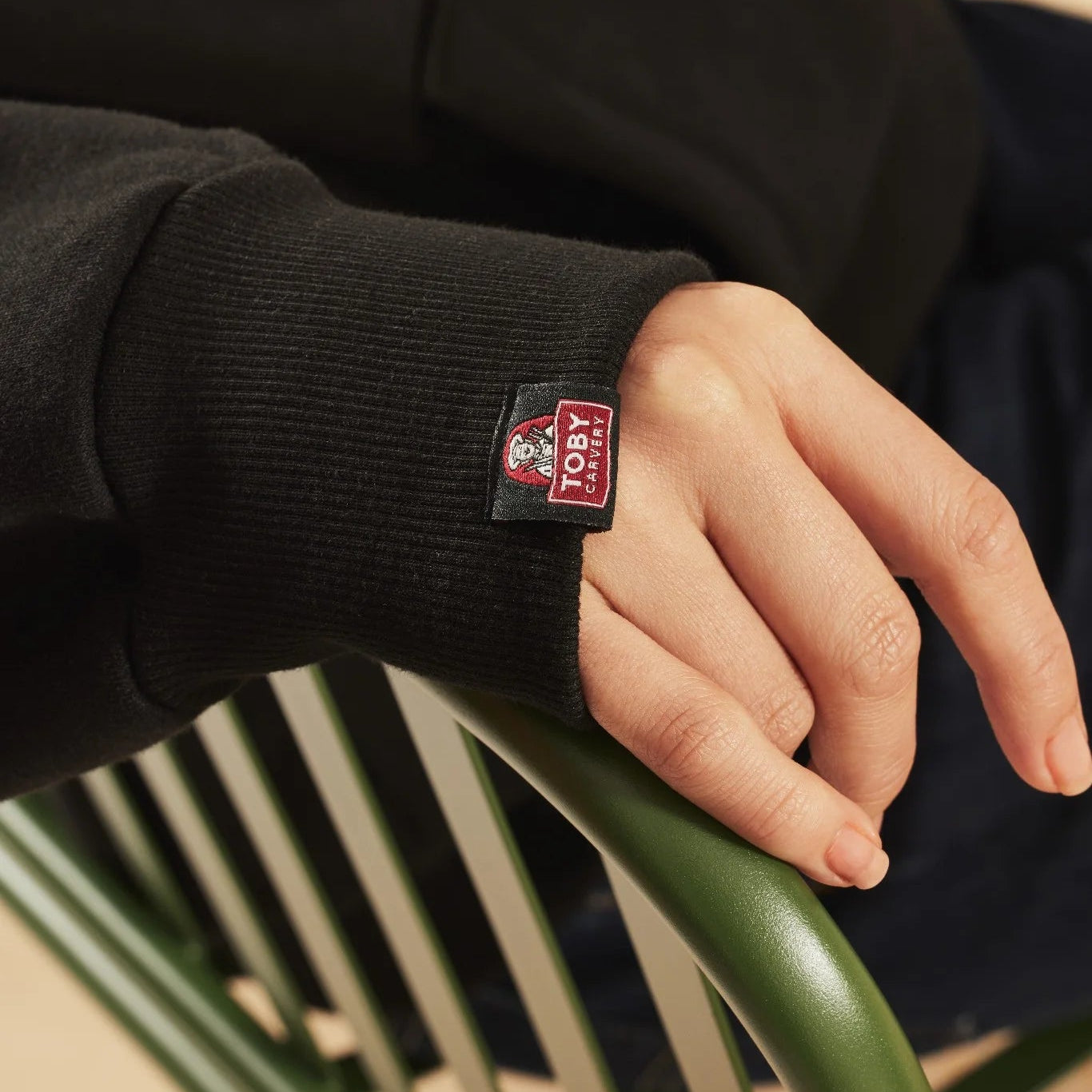 Close-up of a hand wearing a black sweater with a visible Toby Carvery label, sitting on a green chair.