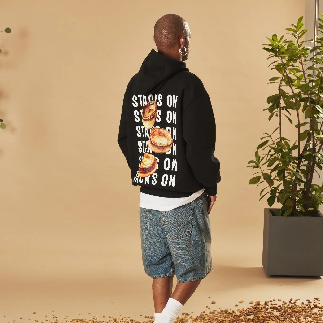 Person wearing a black hoodie with text and graphics, standing against a beige wall with a plant.