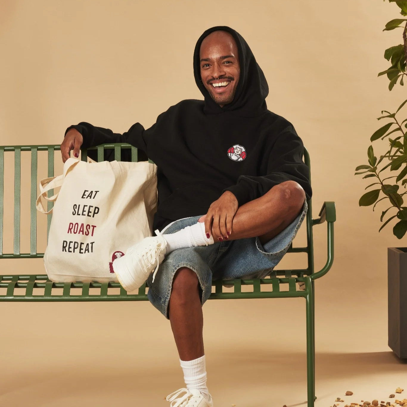 Person sitting on a bench with a tote bag labeled 'Eat Sleep Roast Repeat' against a beige background