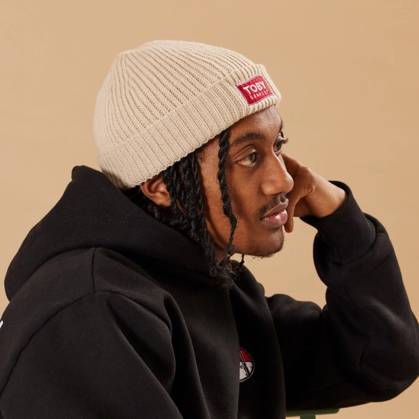 Person wearing a black hoodie and oatmeal beanie with a Toby Carvery logo, sitting against a beige background