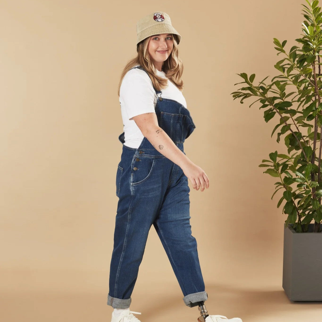 Person wearing blue denim overalls and an oatmeal hat on a beige background with a plant.