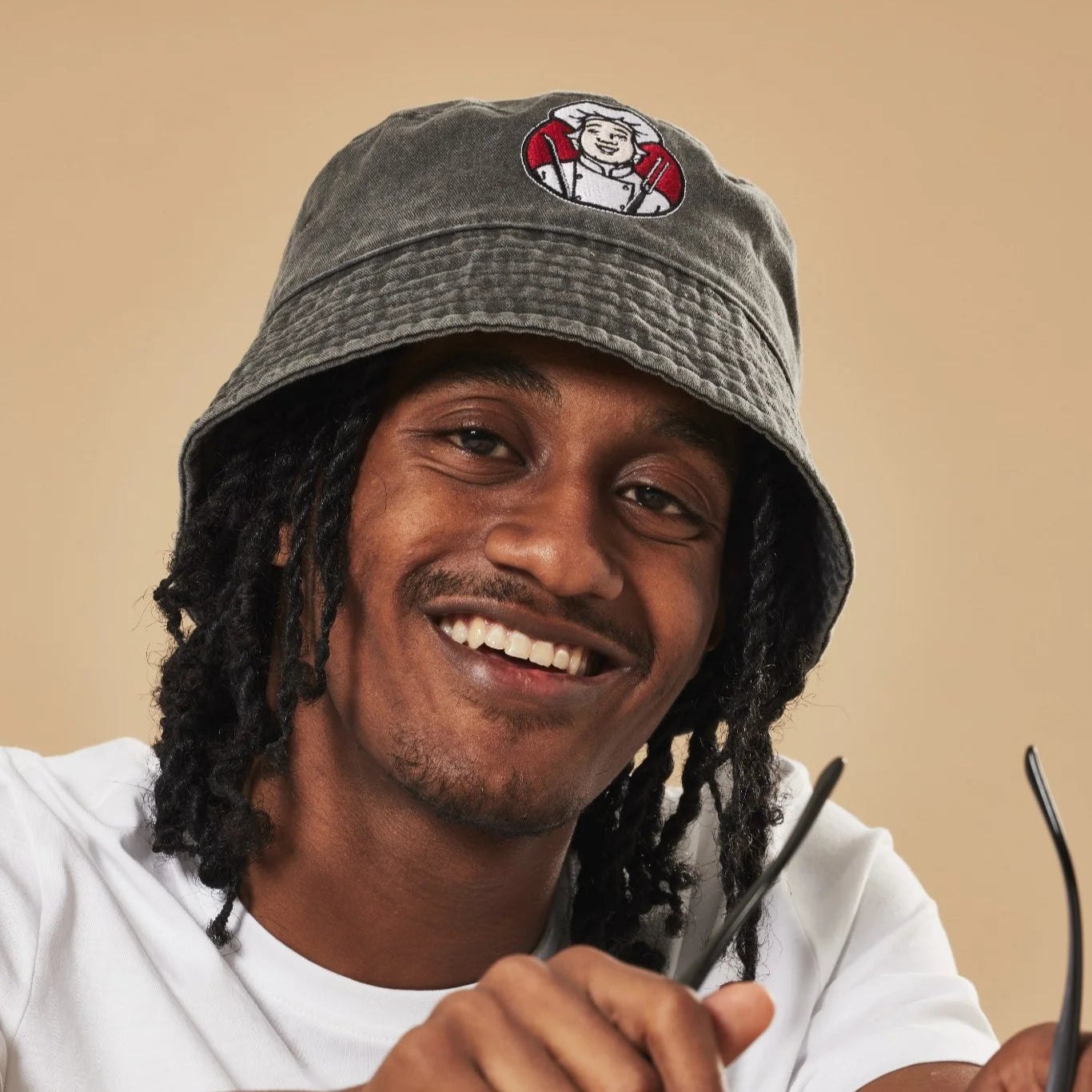 Man wearing a graphite bucket hat with a Toby Carvery logo, holding sunglasses, sitting on a green chair against a beige background.