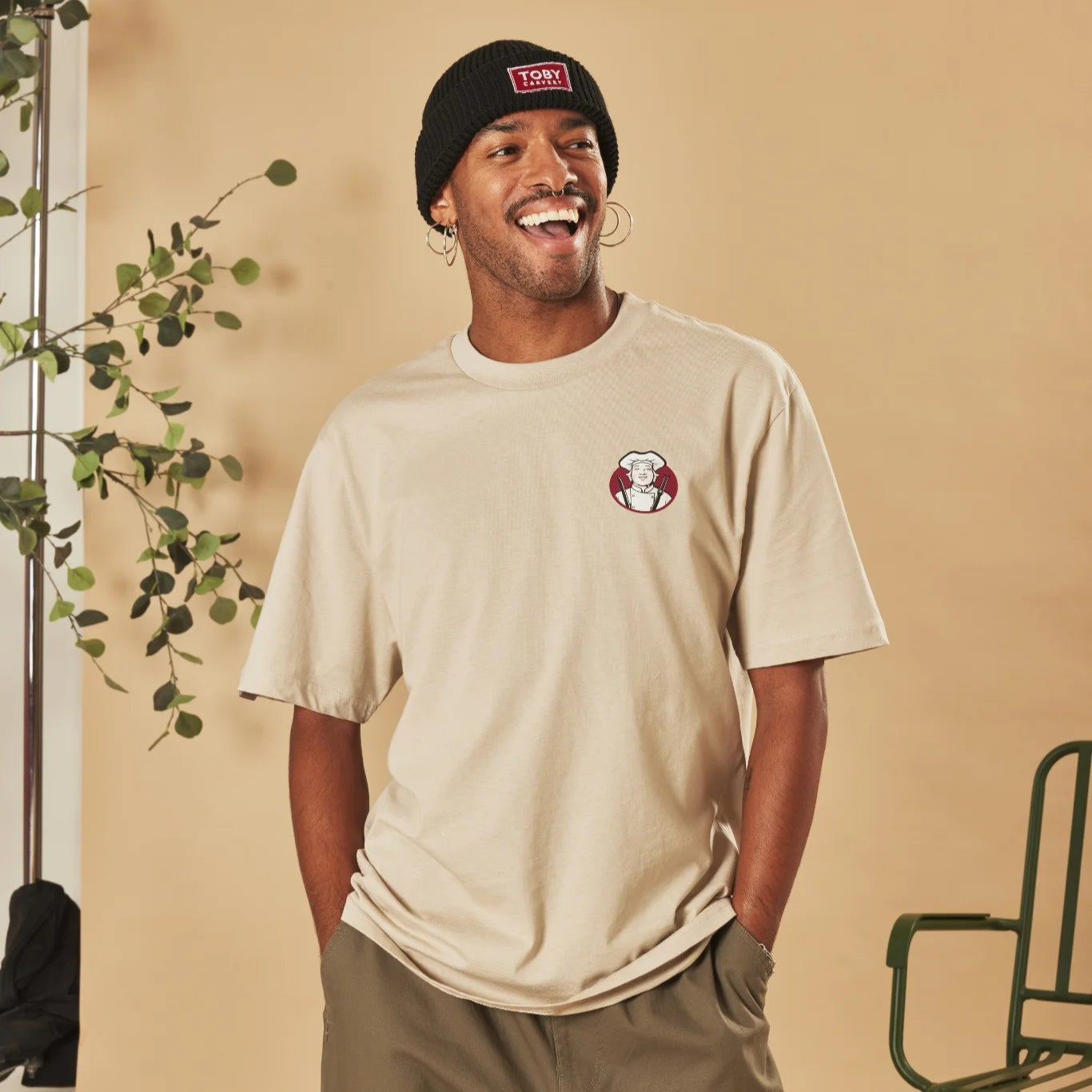 Man wearing a natural stone t-shirt with a Toby Carvery logo and a black beanie, standing against a beige wall with a plant on the left.