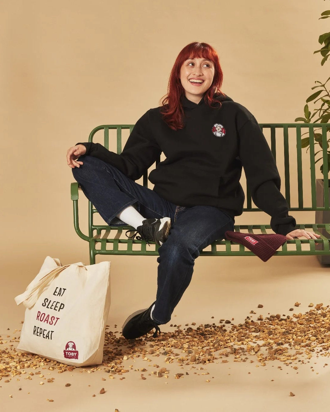 Person sitting on a green bench with plants and a tote bag in a studio setting